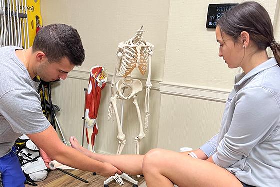 Photo of a young man helping a young woman with an injury on her leg in a medical office