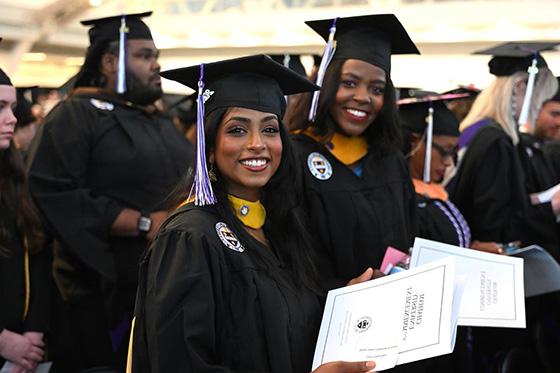 Two graduating students in robes hold up their degree and smile for a photo