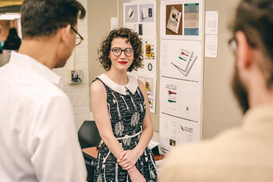 Photo of a female Chatham University student standing in front of her presentation, as two professors offer thoughts.